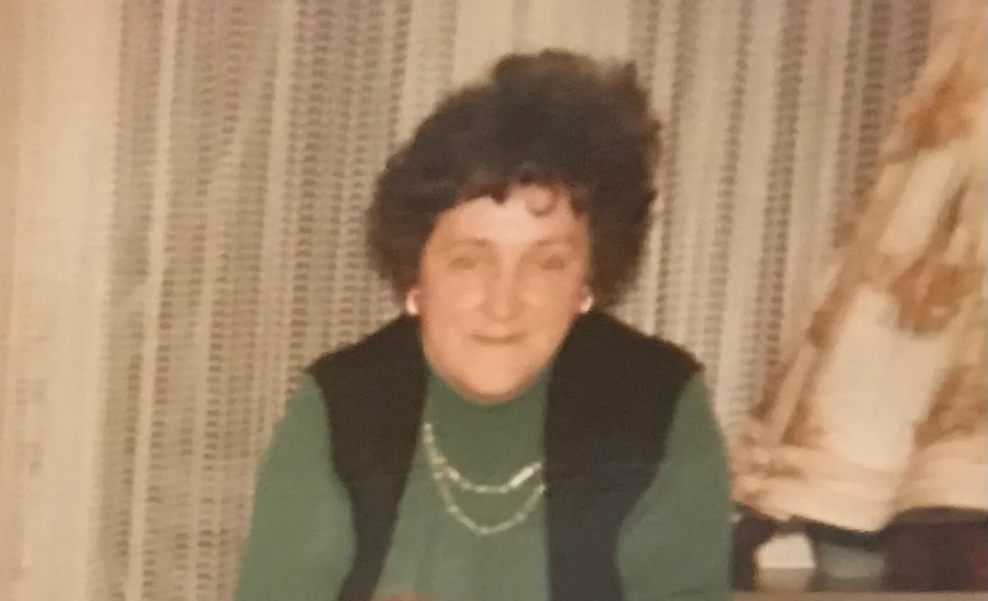 Patricia’s grandma, a white woman with black hair, in front of white curtains. You see her from the chest upwards. She wears a forest green blouse with a black vest on top and a silver necklace and white earrings.