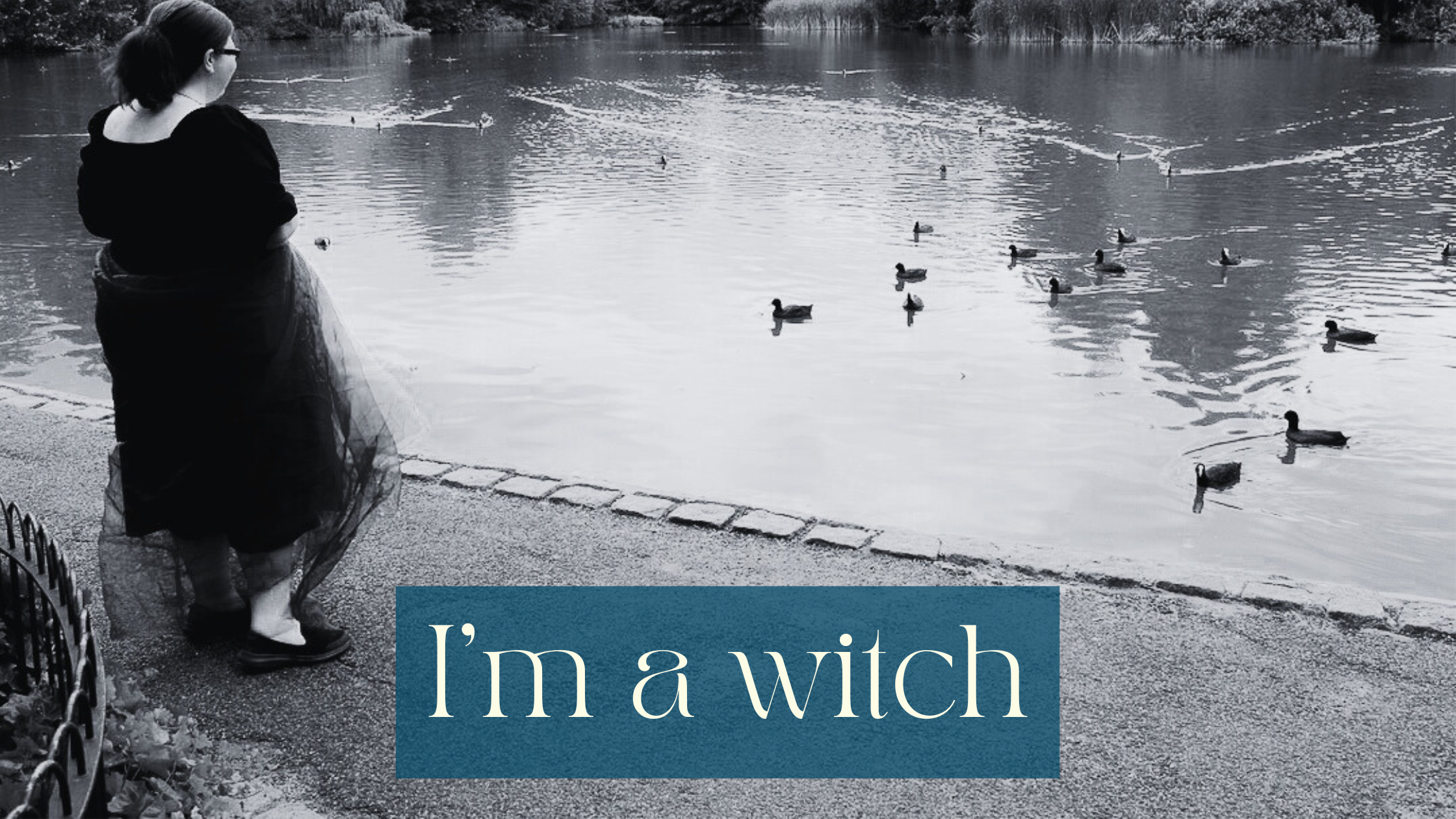 Patricia, a fat white woman with glasses and her hair tied up, standing on the left in front of a lake. She has a veil wrapped around her waist, her back is to the viewer and all the ducks in the lake are moving towards her. The photo is black and white. White writing on a half transparent petrol coloured background: I’m a witch
