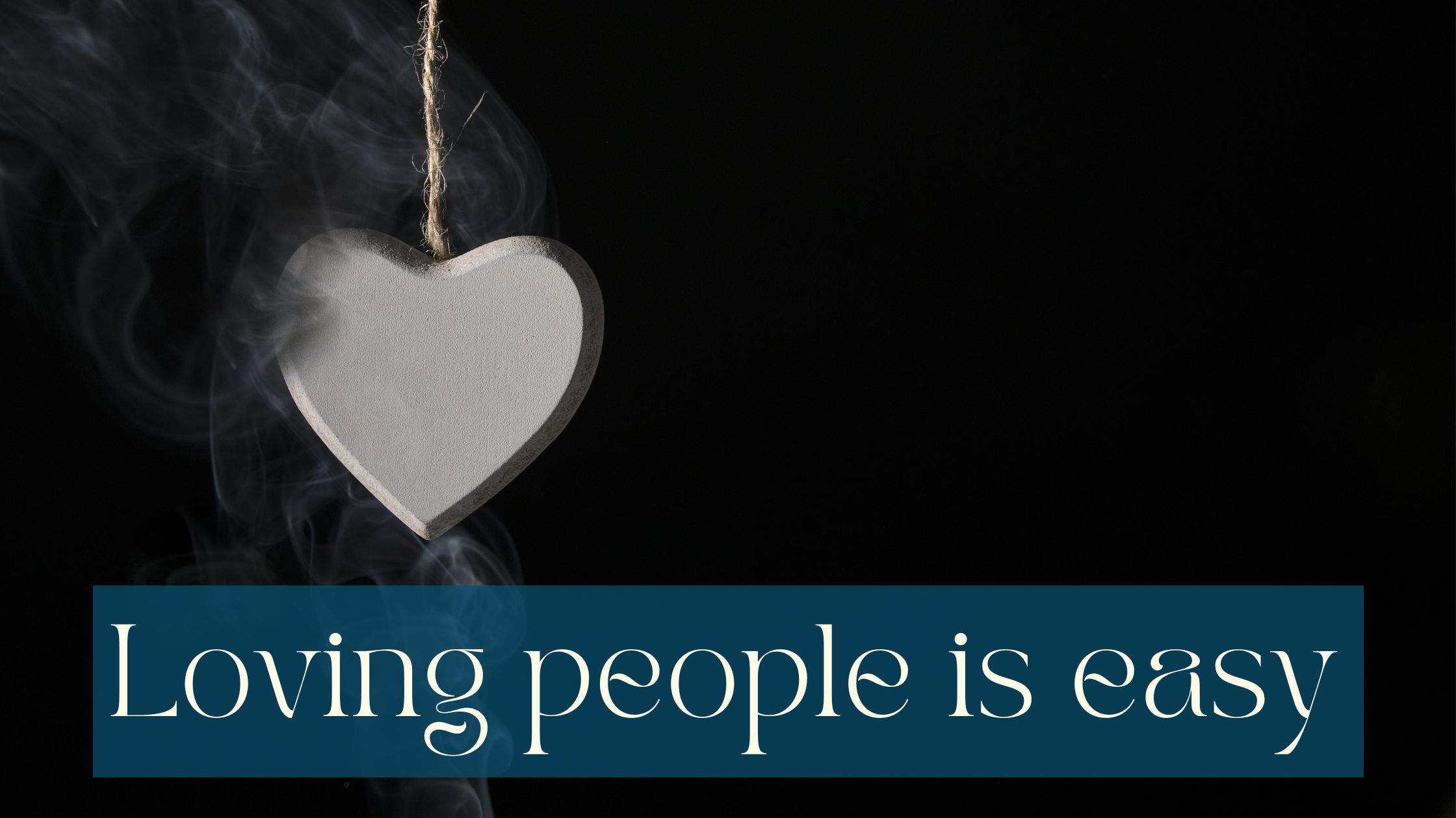 Black background with a white-ish wooden heart on a porous string on it. Smoke is coming from below, touching the heart. It seems to come from the heart at the same time, as if it was burning. White writing on slightly transparent petrol background: Loving people is easy.